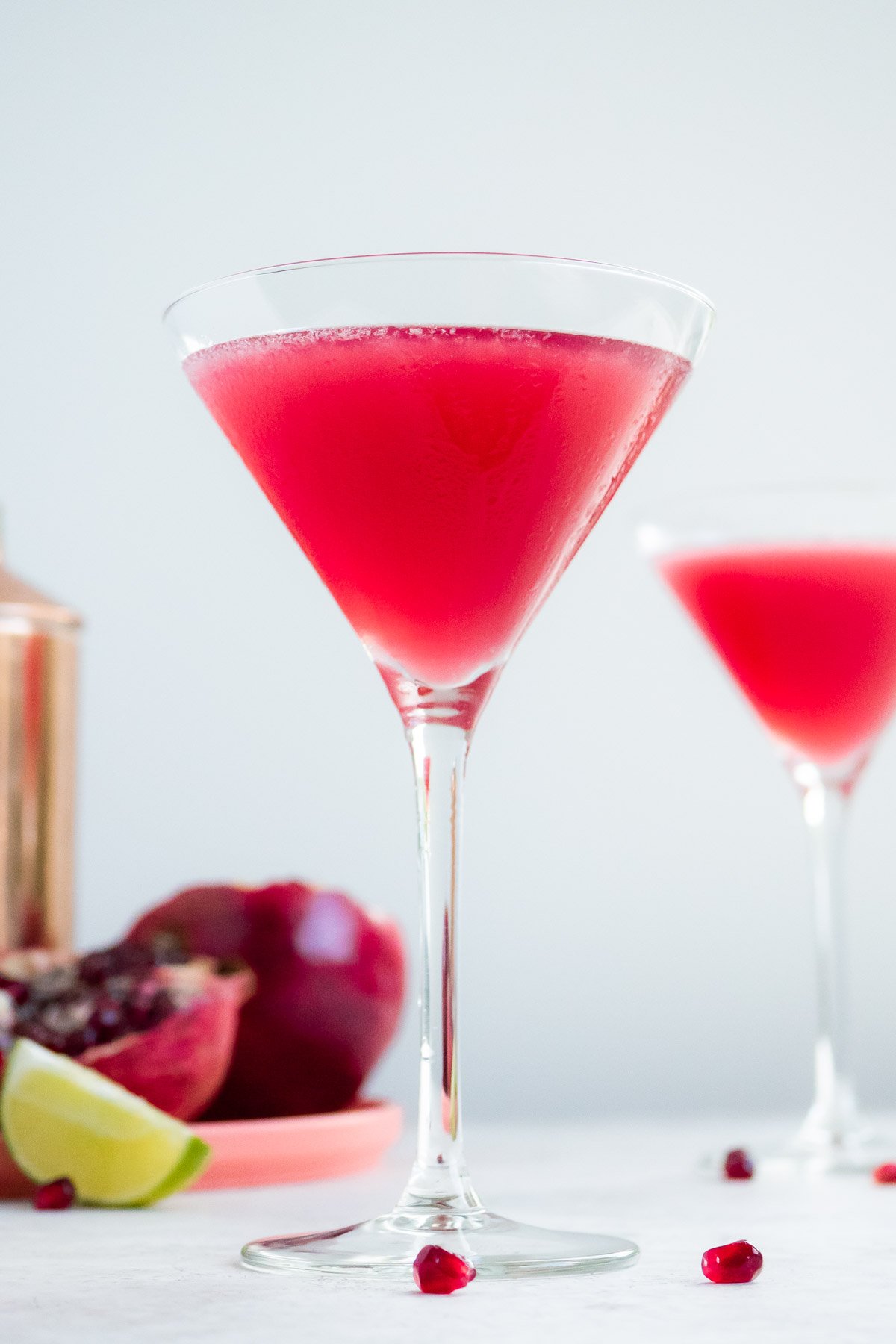 a pomegranate martini in a tall martini glass with some fruit and another martini in the background.