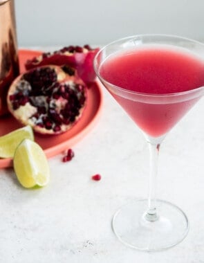 a pomegranate martini in a tall martini glass with a martini shaker, two halve of a pomegranate and two lime wedges in the background.