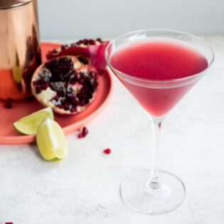 a pomegranate martini in a tall martini glass with a martini shaker, two halve of a pomegranate and two lime wedges in the background.