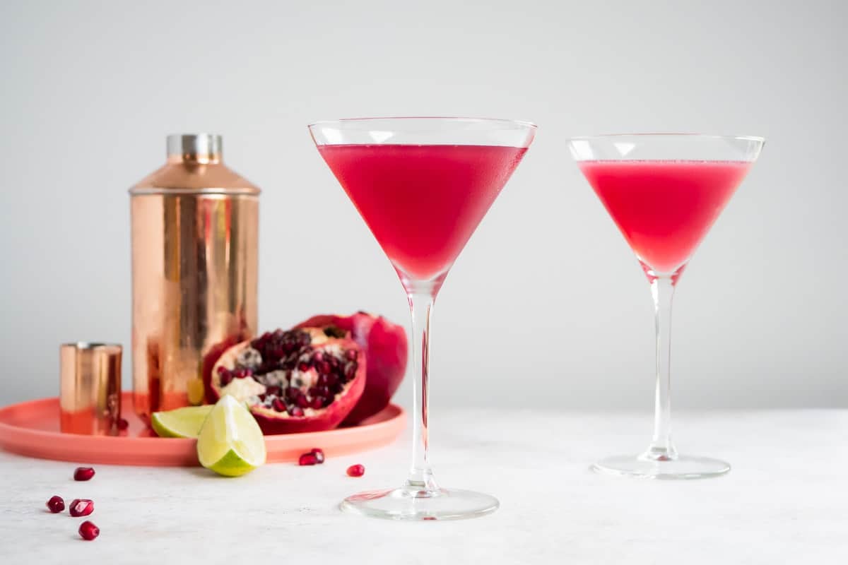 a pomegranate martini in a tall martini glass with another martini, martini shaker, half of a pomegranate and 2 lime wedges in the background.