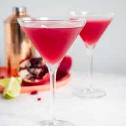 a pomegranate martini in a tall martini glass with another martini, martini shaker, half of a pomegranate and 2 lime wedges in the background.