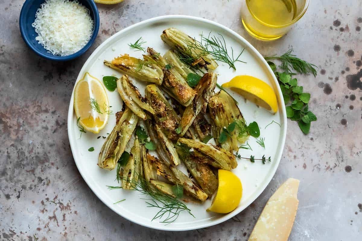 roasted fennel with parmigiano cheese with three lemon wedges on a plate next to a parmigiano-reggiano cheese rind, a cup of olive oil, and a bowl of parmigiano-reggiano cheese.