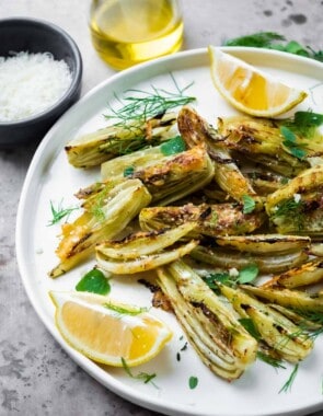 roasted fennel with parmigiano cheese with two lemon wedges on a plate next to a cup of olive oil, and a bowl of parmigiano-reggiano cheese.