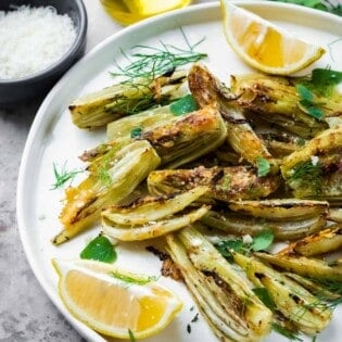 roasted fennel with parmigiano cheese with two lemon wedges on a plate next to a cup of olive oil, and a bowl of parmigiano-reggiano cheese.