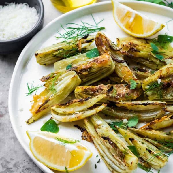 roasted fennel with parmigiano cheese with two lemon wedges on a plate next to a cup of olive oil, and a bowl of parmigiano-reggiano cheese.