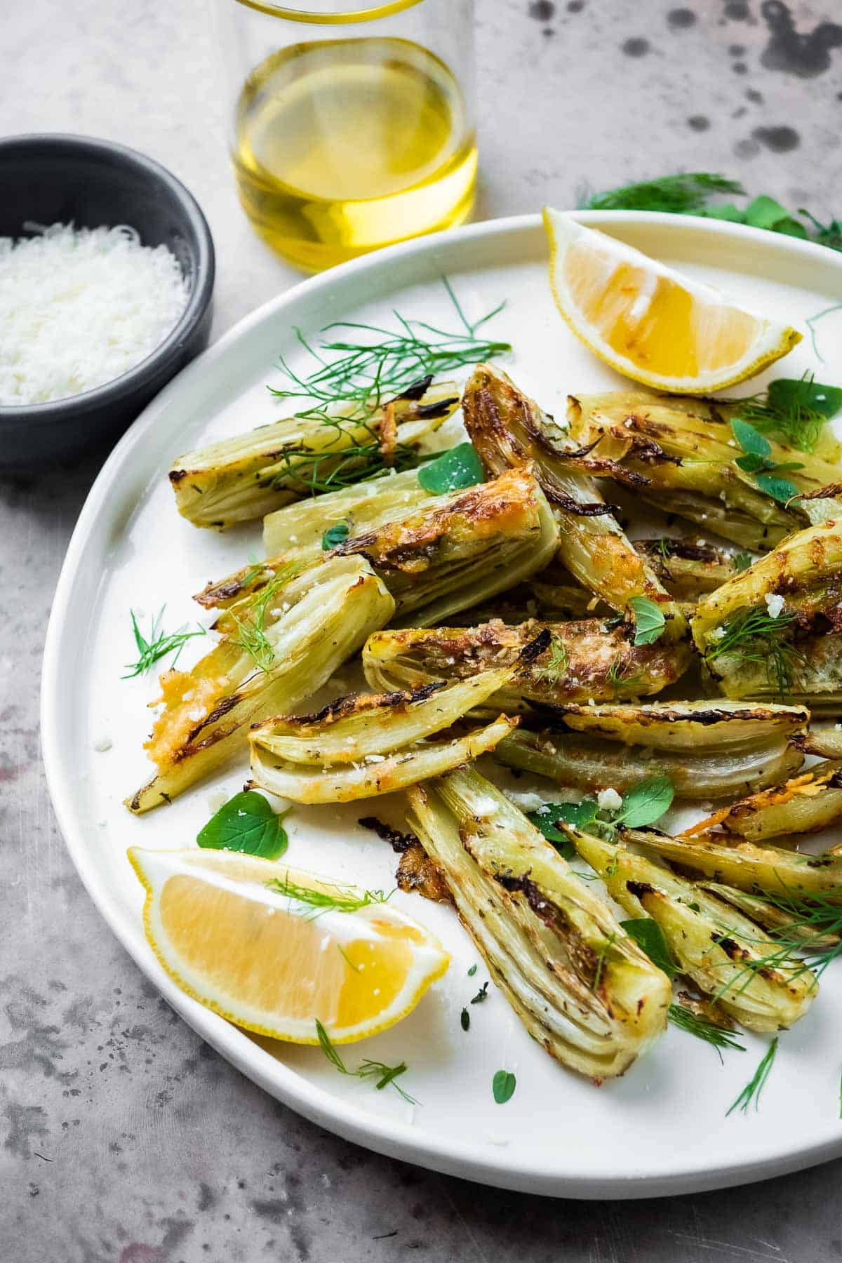 roasted fennel with parmigiano cheese with two lemon wedges on a plate next to a cup of olive oil, and a bowl of parmigiano-reggiano cheese.