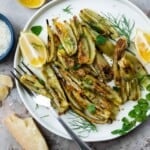 roasted fennel with parmigiano cheese with two lemon wedges on a plate next to another lemon wedge, a parmigiano-reggiano cheese rind and a bowl of parmigiano-reggiano cheese.