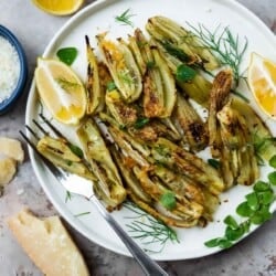 roasted fennel with parmigiano cheese with two lemon wedges on a plate next to another lemon wedge, a parmigiano-reggiano cheese rind and a bowl of parmigiano-reggiano cheese.