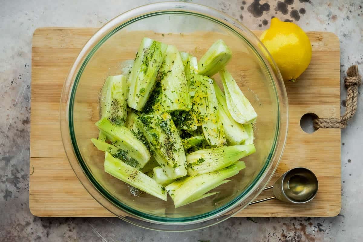 pieces of seasoned uncooked fennel in bowl next to a measuring spoon and a lemon on a cutting board.