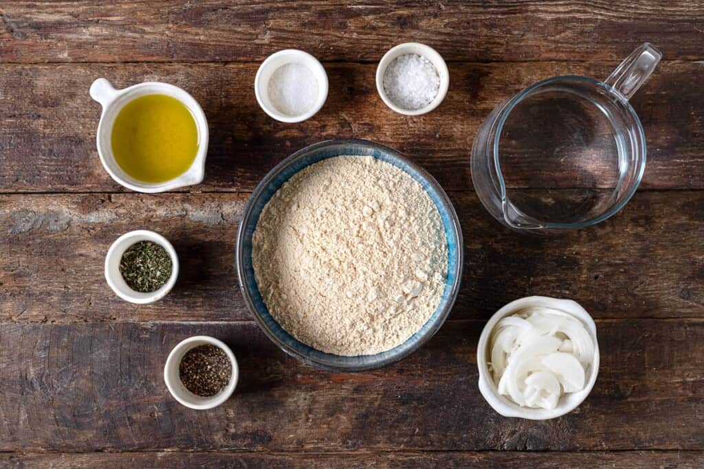 ingredients for a farinata italian chickpea pancake including chickpea flour, fine sea salt, flaky sea salt, olive oil, water, rosemary, sliced onion and pepper.