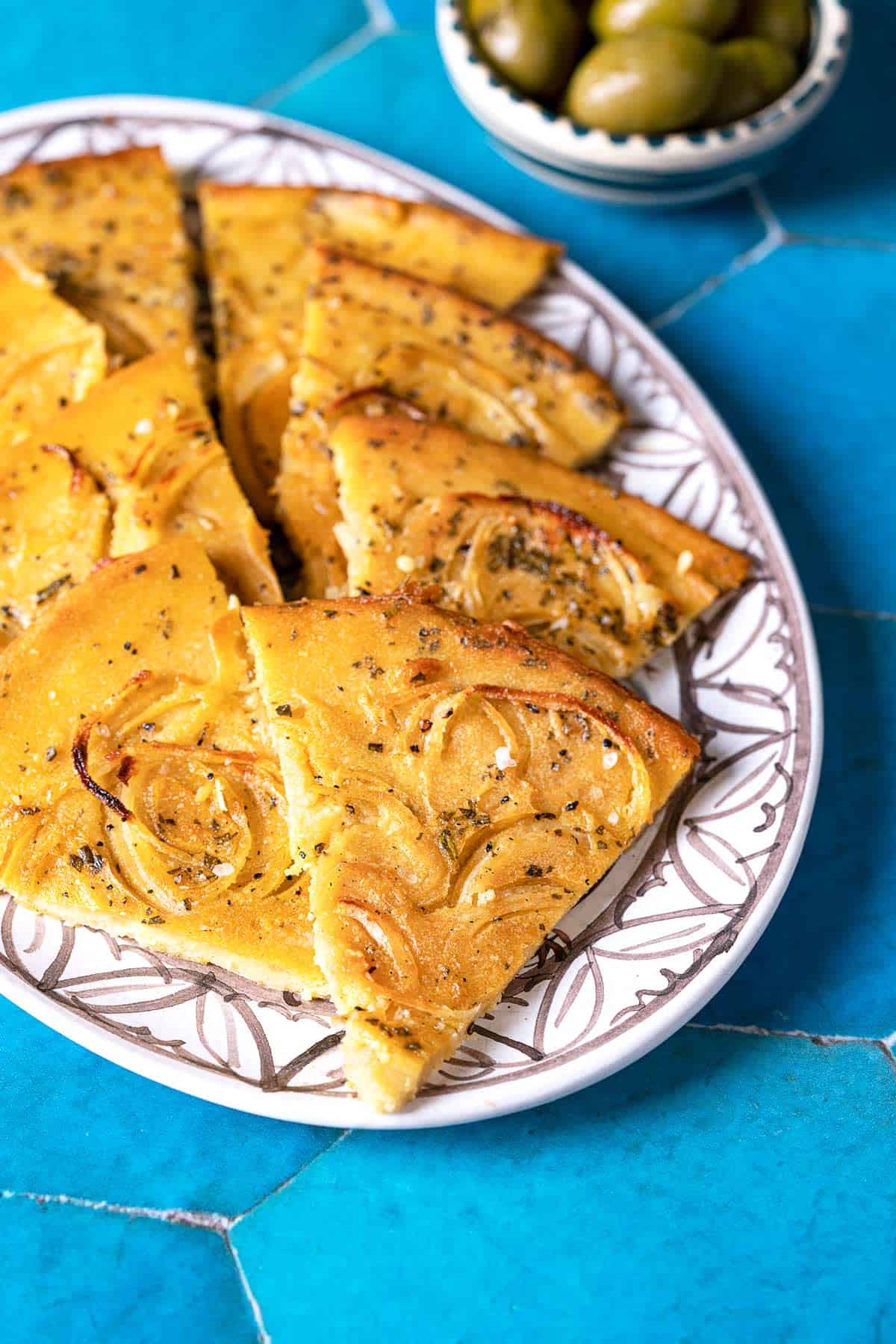 slices of a baked farinata italian chickpea pancake on a plate next to a bowl of green olives.