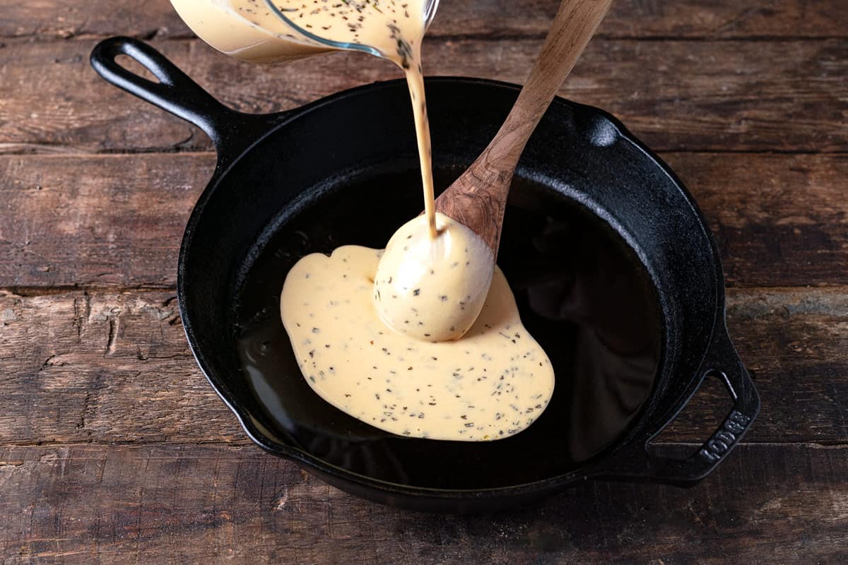 farinata italian chickpea pancake batter being poured into a cast iron skillet over a wooden spoon.