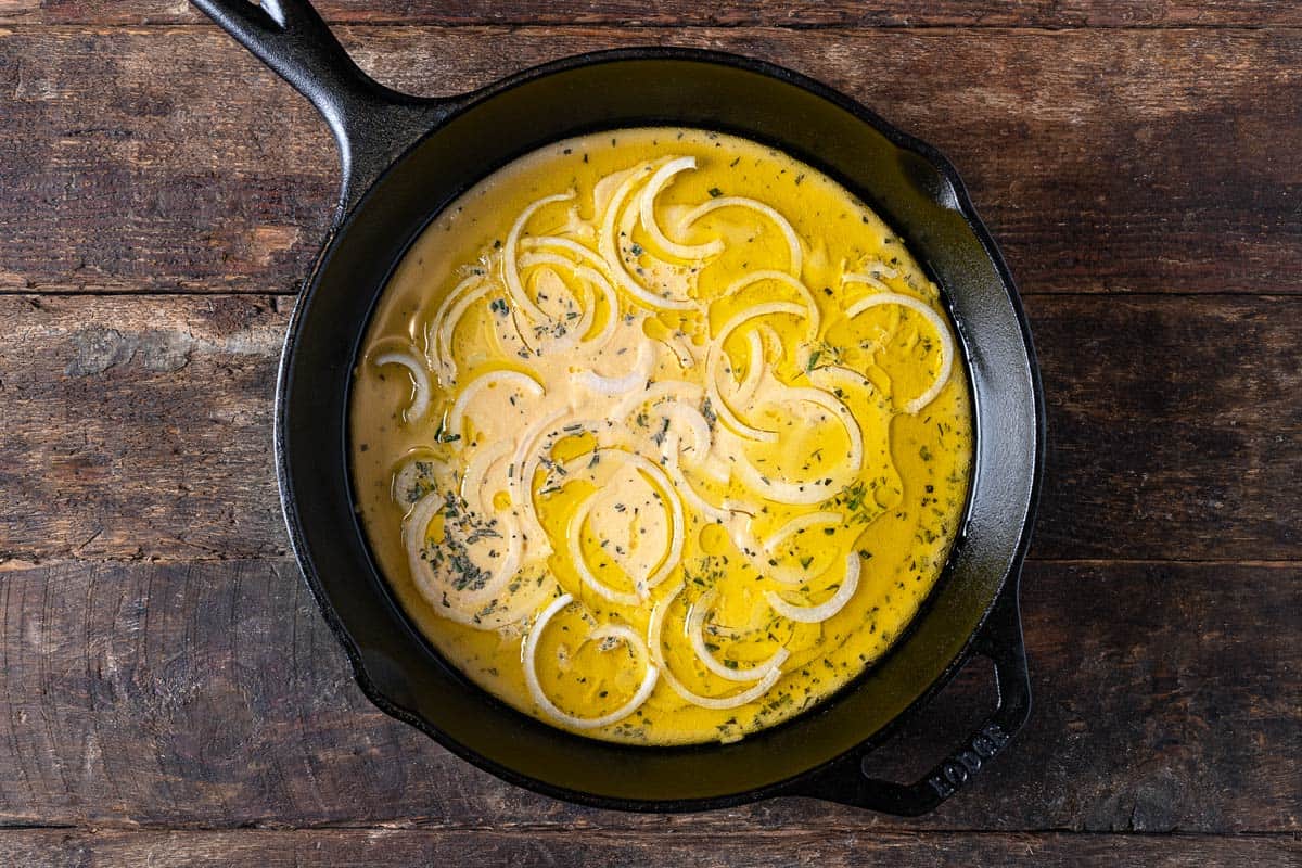 unbaked farinata italian chickpea pancake batter with sliced onions in a cast iron skillet.