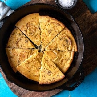 a baked farinata italian chickpea pancake sliced into pieces in a cast iron skillet next to a bowl of flaky sea salt.