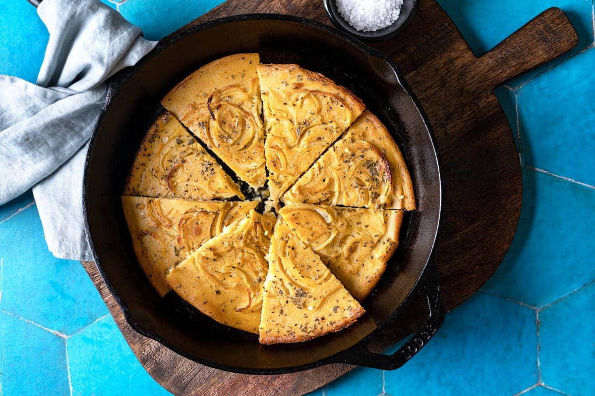 a baked farinata italian chickpea pancake sliced into pieces in a cast iron skillet next to a bowl of flaky sea salt.