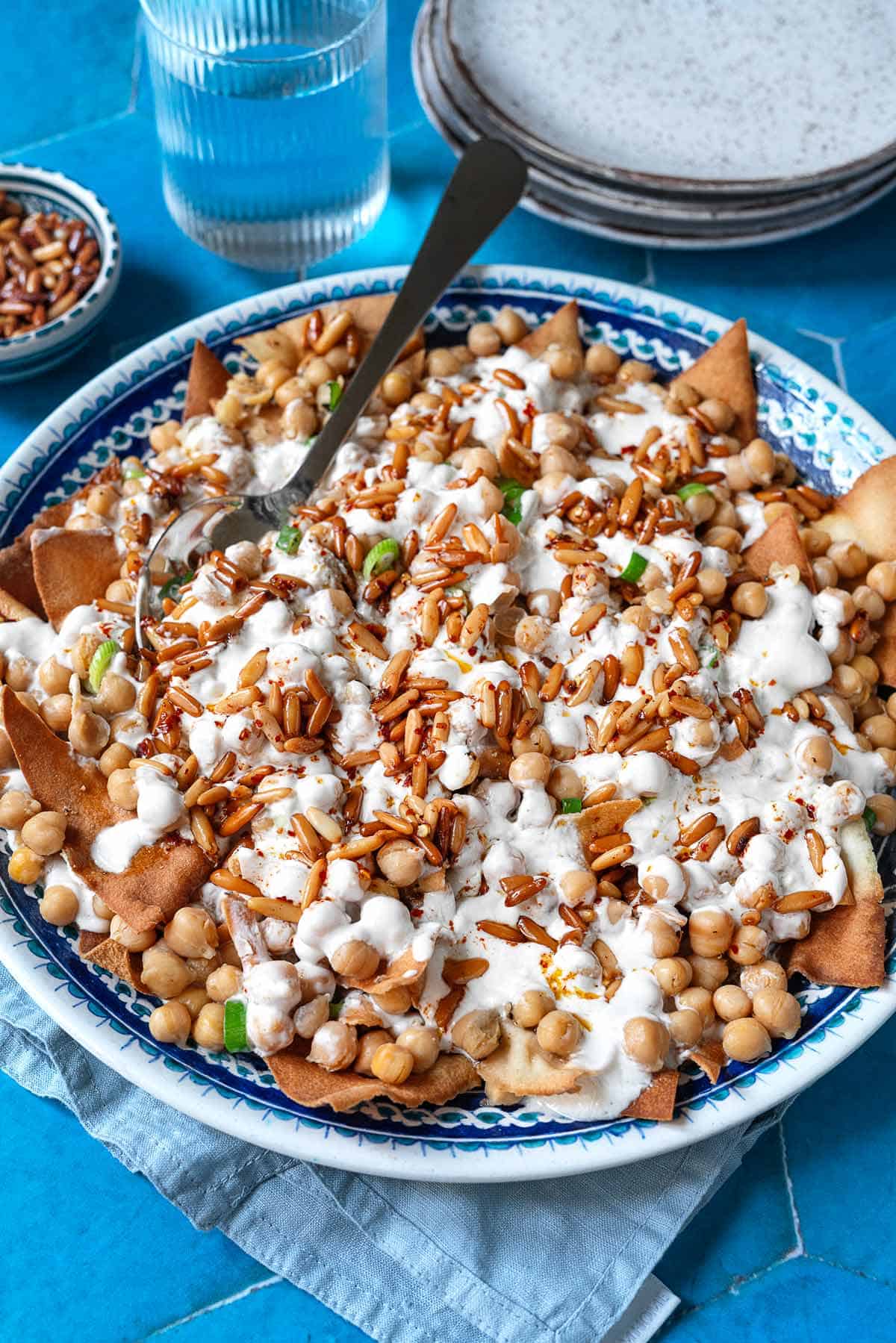 Fatteh (Spiced Chickpeas with Crispy Pita and Garlicky Yogurt) in a bowl with a spoon next to a glass of water.