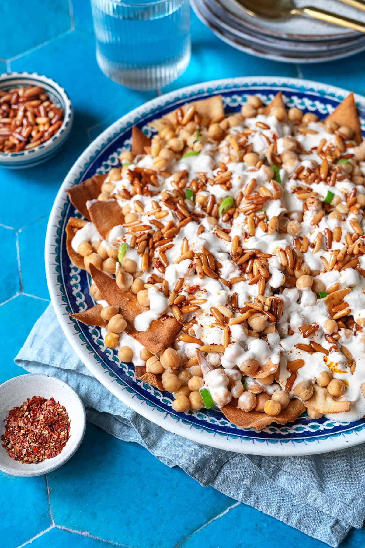 Fatteh (Spiced Chickpeas with Crispy Pita and Garlicky Yogurt) in a bowl next to a bowl of pine nuts and a bowl of spices.
