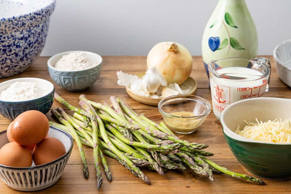 Ingredients for asparagus quiche, including eggs, asparagus, grated cheese, milk, flour, onion, garlic, and Dijon mustard.