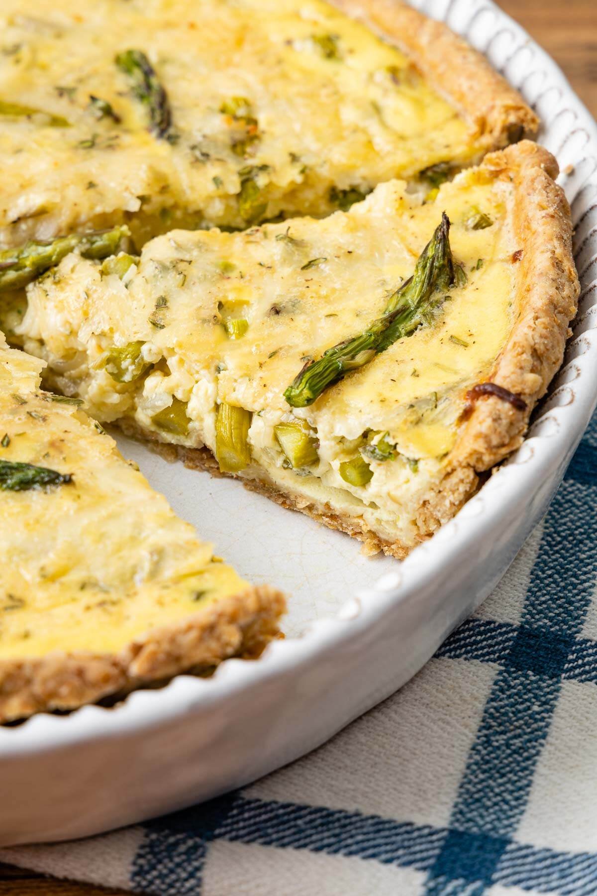 Side shot of sliced asparagus quiche in the tin with a blue and white checked background.