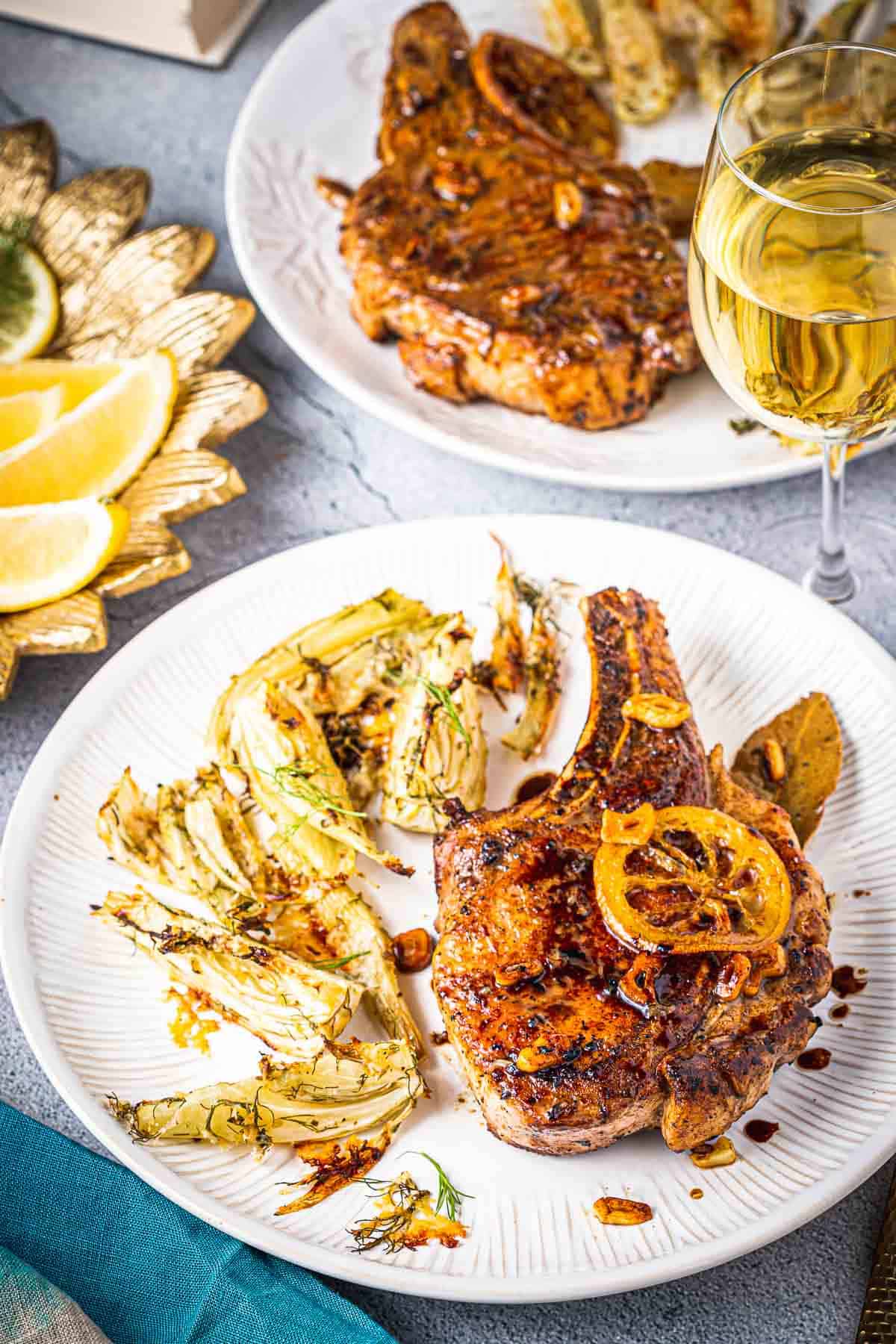 2 plates with pan seared pork chops on a plate with roasted fennel next to a glass of wine.