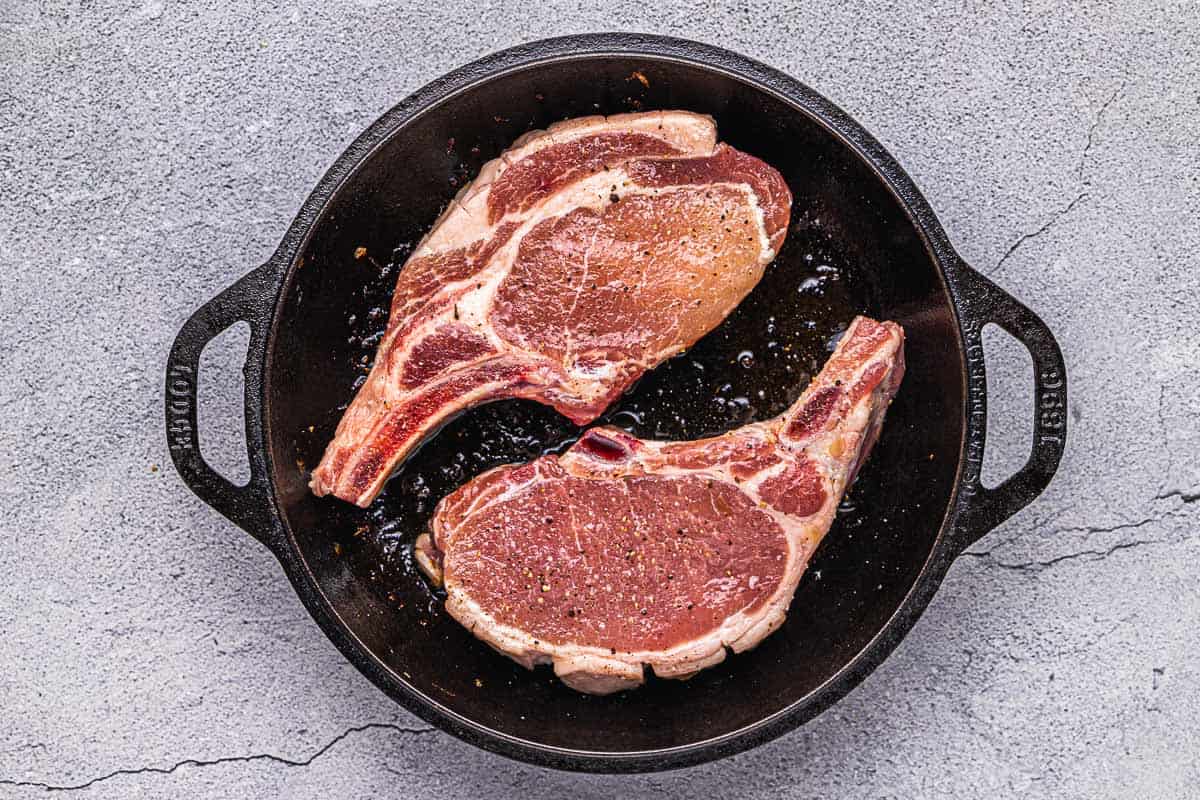 two pork chops seasoned with salt and pepper cooking in a cast iron skillet.