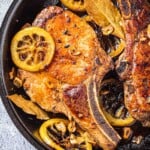 close up of two pan seared pork chops in a cast iron skillet with bay leaves, sliced garlic and fried lemon rings.