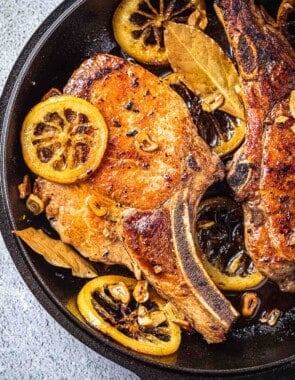 close up of two pan seared pork chops in a cast iron skillet with bay leaves, sliced garlic and fried lemon rings.