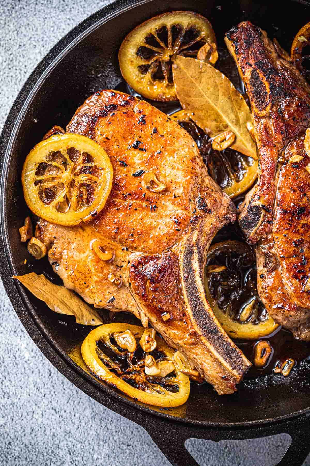 close up of two pan seared pork chops in a cast iron skillet with bay leaves, sliced garlic and fried lemon rings.