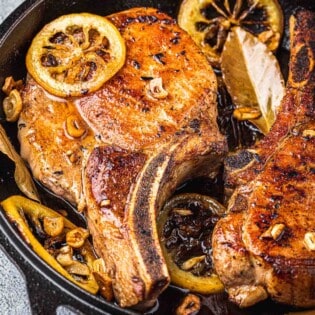 close up of two pan seared pork chops in a cast iron skillet with bay leaves and fried lemon rings.