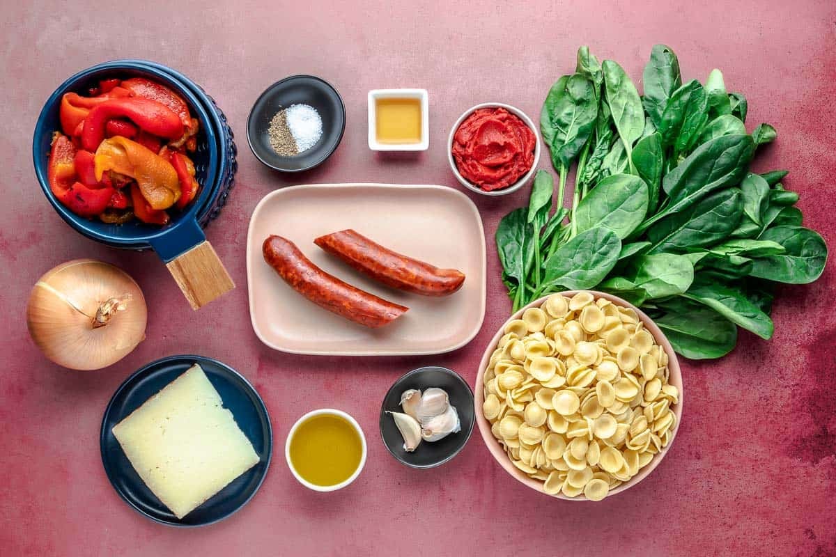 Ingredients for Chorizo Pasta, including extra virgin olive oil, Spanish chorizo, onion, garlic, salt, tomato paste, roasted red peppers, spinach, vinegar, black pepper, manchego cheese, and short pasta.