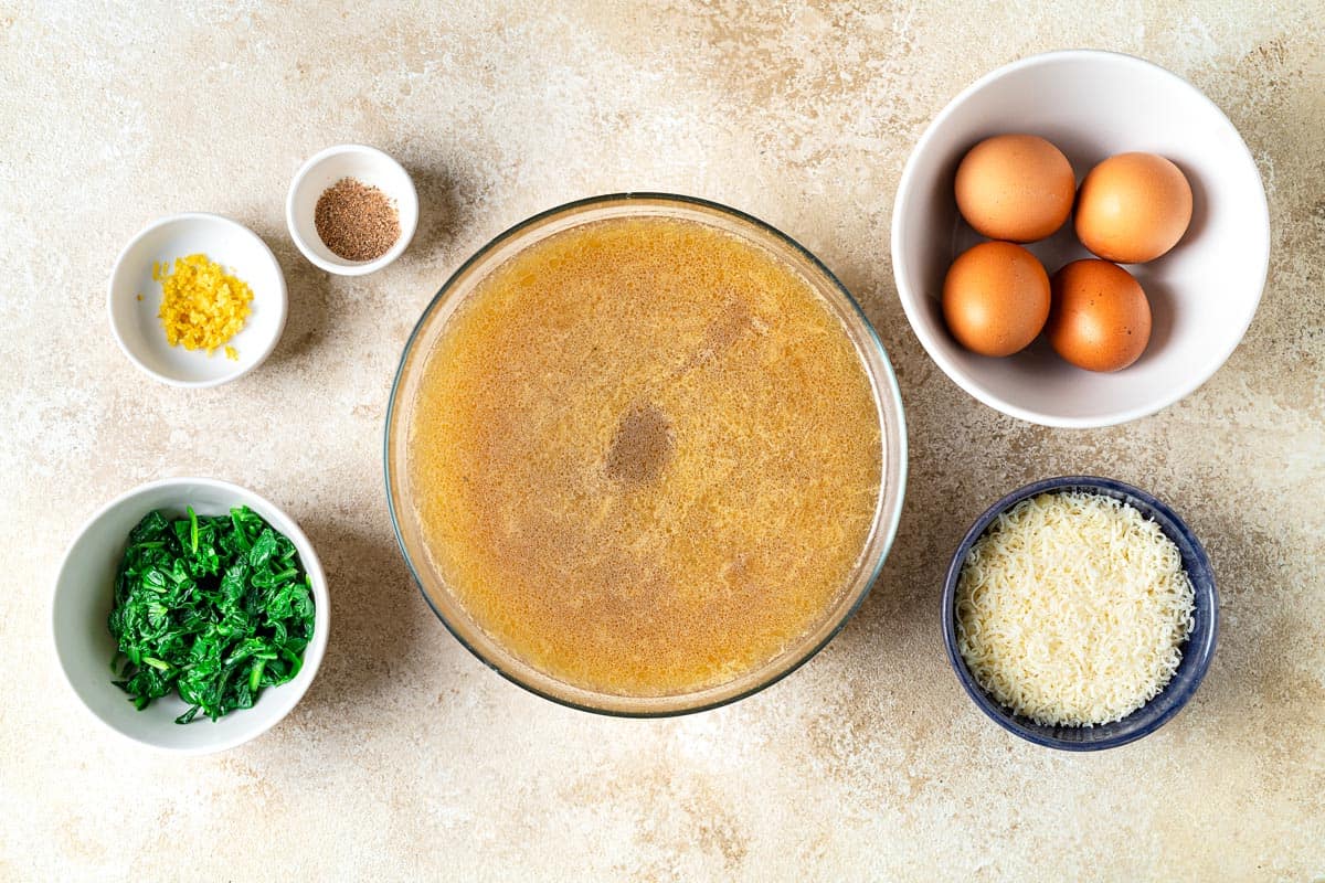 Overhead shot showing ingredients for Stracciatella Soup, including chicken broth, eggs, grated nutmeg, lemon zest, and wilted spinach.