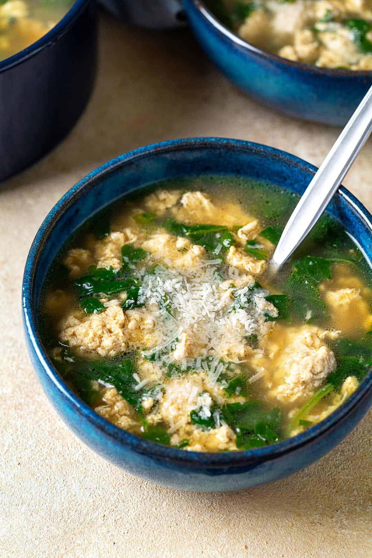 Close up of a blue bowl with Stracciatella Soup and freshly grated Parmesan cheese.