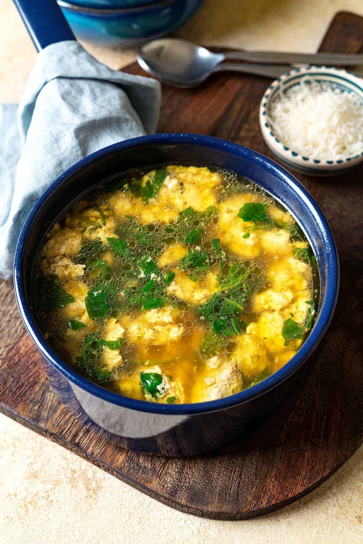 Blue pot with Stracciatella Soup and freshly grated Parmesan cheese on the side.