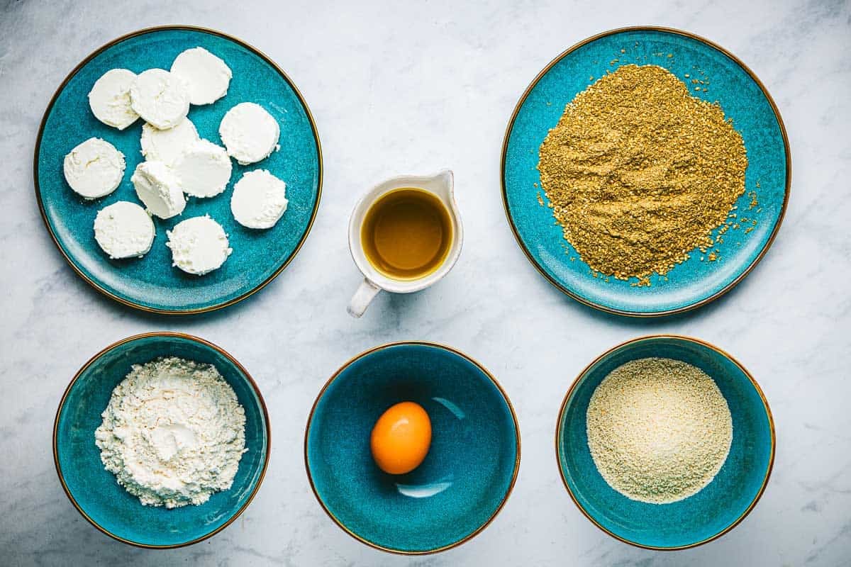 ingredients for fried goat cheese slices including goat cheese, egg, flour, bread crumbs, olive oil and za'atar.