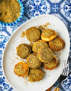 10 fried goat cheese slices on plate next to a bowl of za'ata and a fork.