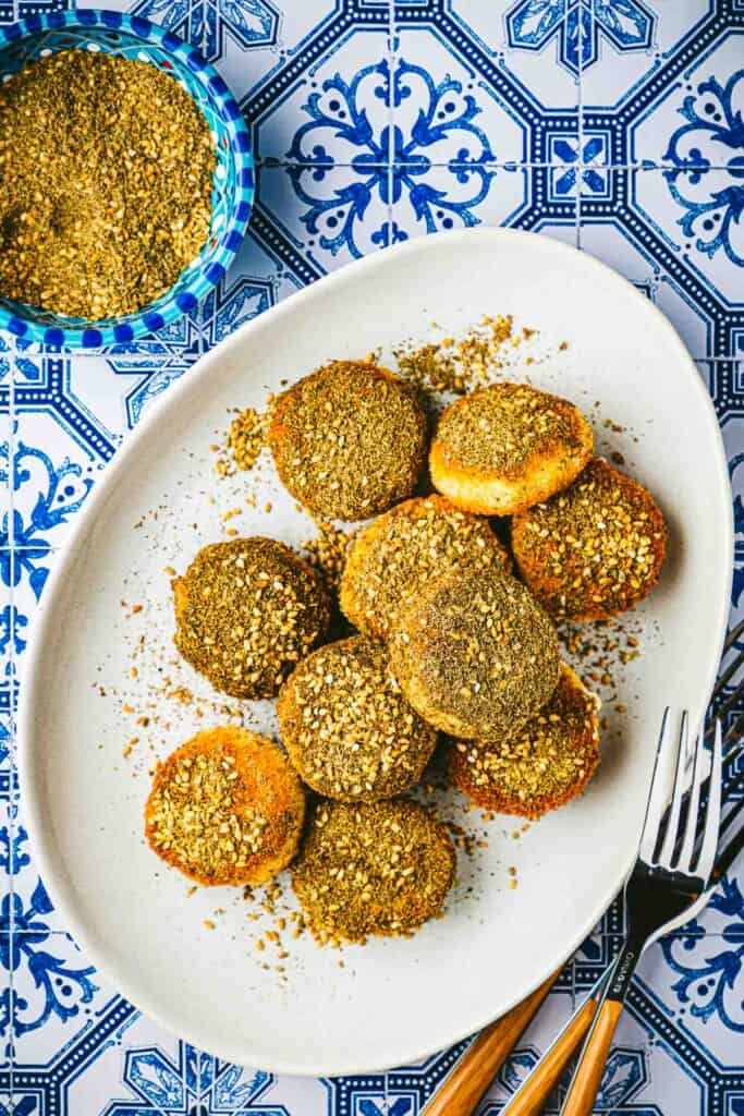 10 fried goat cheese slices on plate next to a bowl of za'ata and a fork.