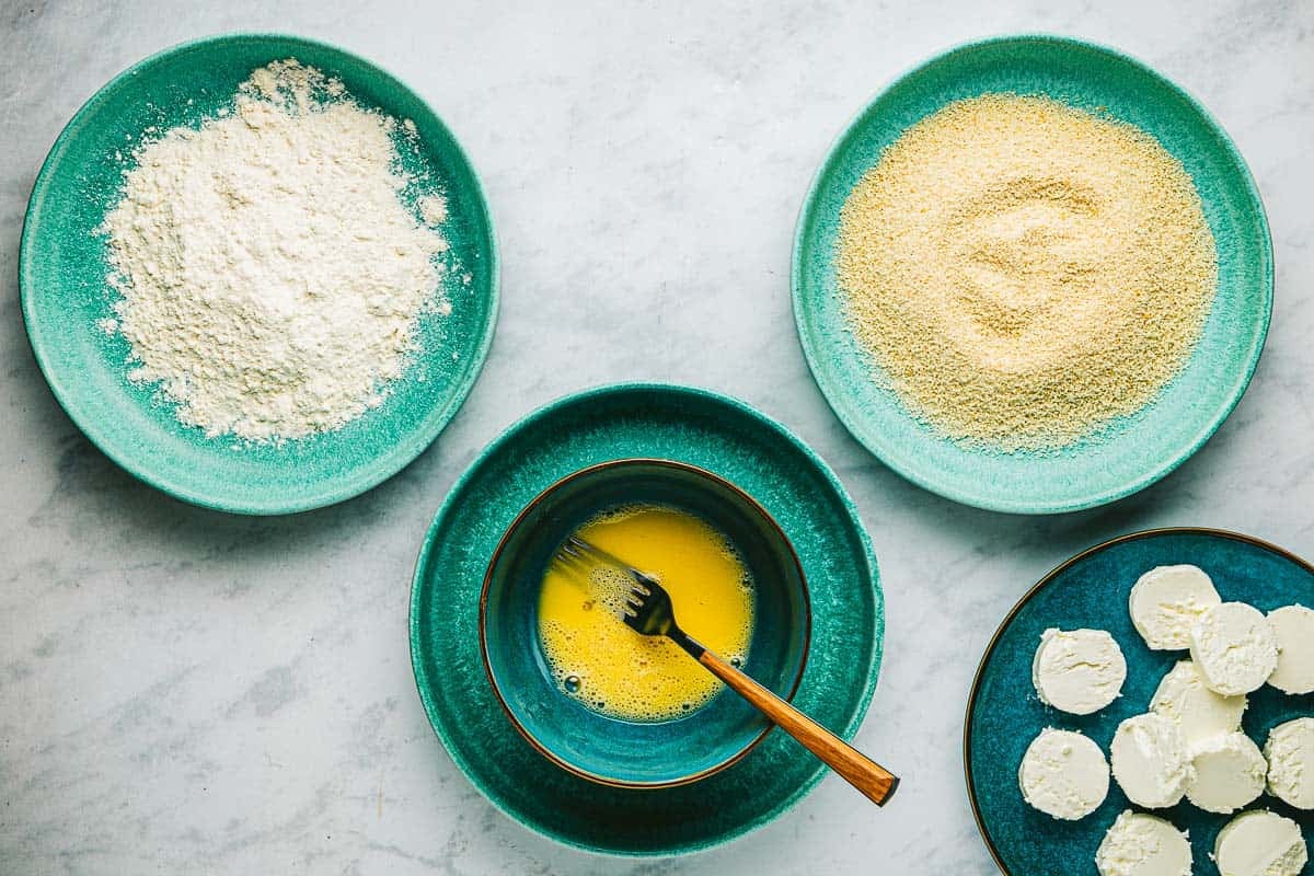 ingredients for dredging including goat cheese slices, egg in a bowl with a fork, a plate of flour and a plate of bread crumbs.