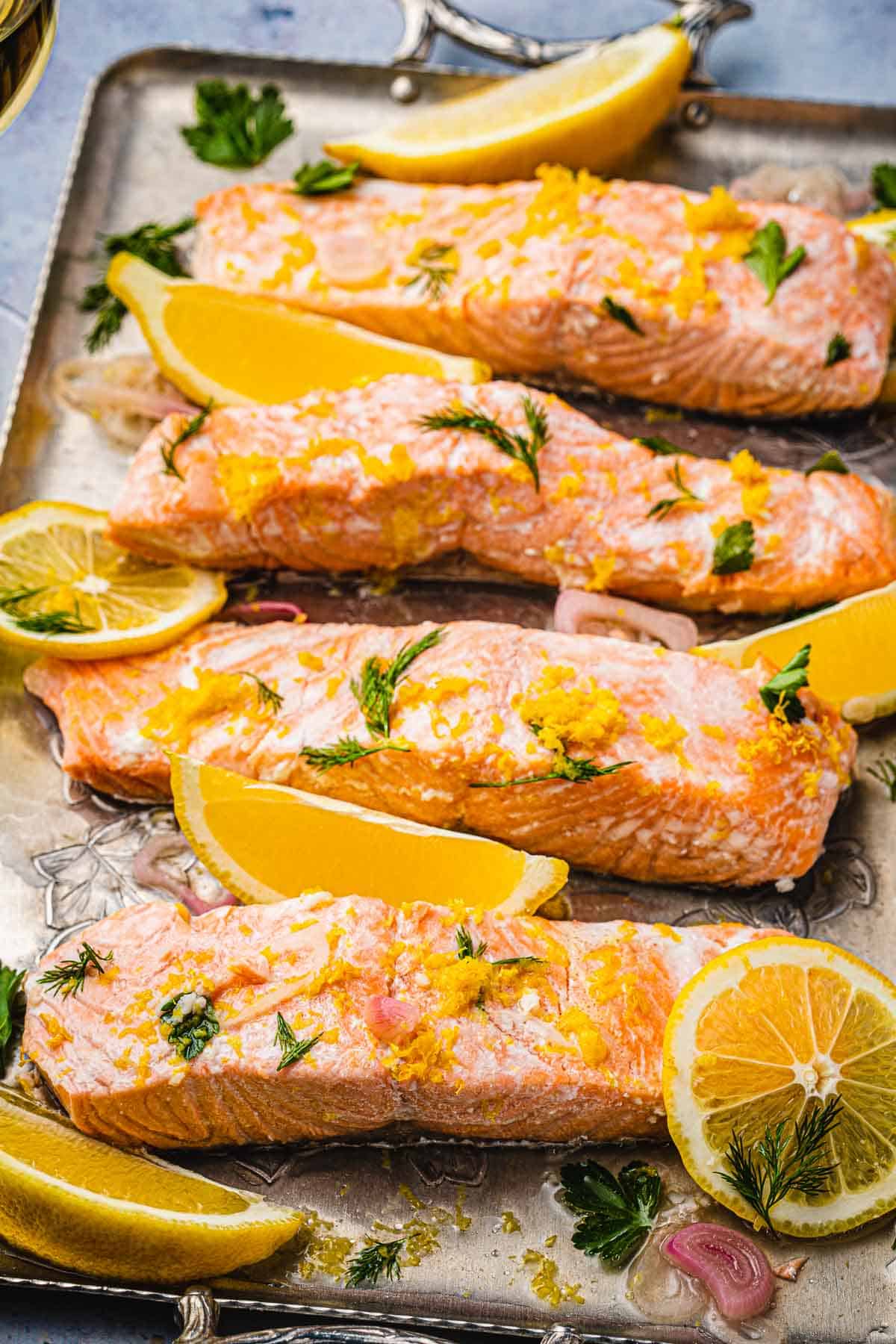 4 poached salmon fillet slices with dill, lemon, parsley and shallots on a serving tray.