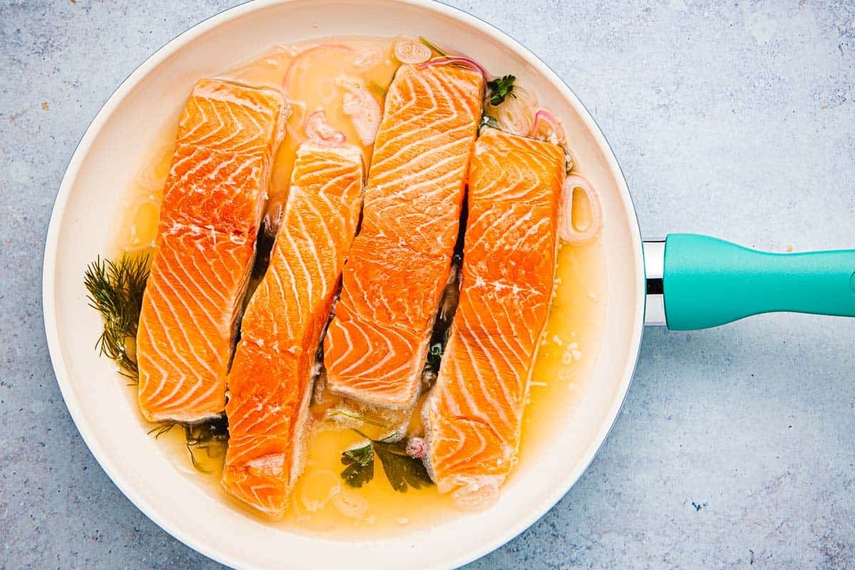 4 uncooked salmon fillets being poached in a skillet.