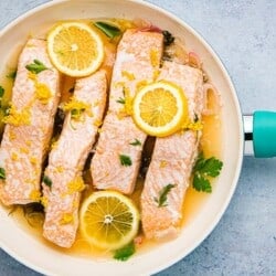Poached salmon in a white skillet with a blue handle. The salmon also has lemon slices and fresh herbs scattered on top.