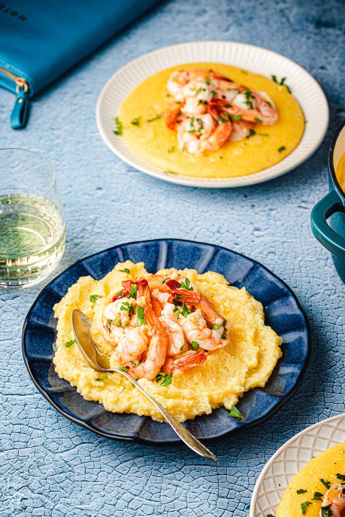 two plates of polenta and shrimp garnished with parsley next to a glass of wine.