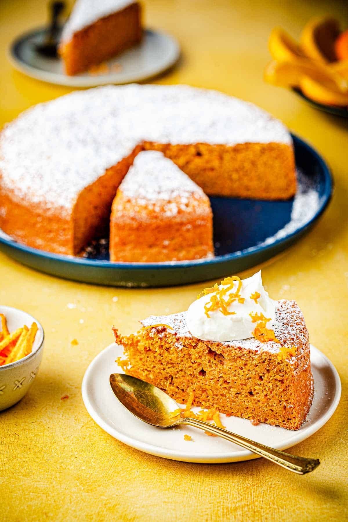 A slice of Torta di Carote (Italian Carrot Cake) topped with a dollop of Greek yogurt, a drizzle of honey and orange zest on a plate with a spoon.