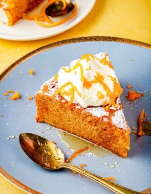 A slice of Torta di Carote (Italian Carrot Cake) topped with a dollop of Greek yogurt, a drizzle of honey and orange zest on a plate with a spoon.