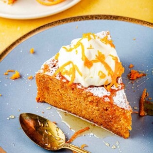 A slice of Torta di Carote (Italian Carrot Cake) topped with a dollop of Greek yogurt, a drizzle of honey and orange zest on a plate with a spoon.