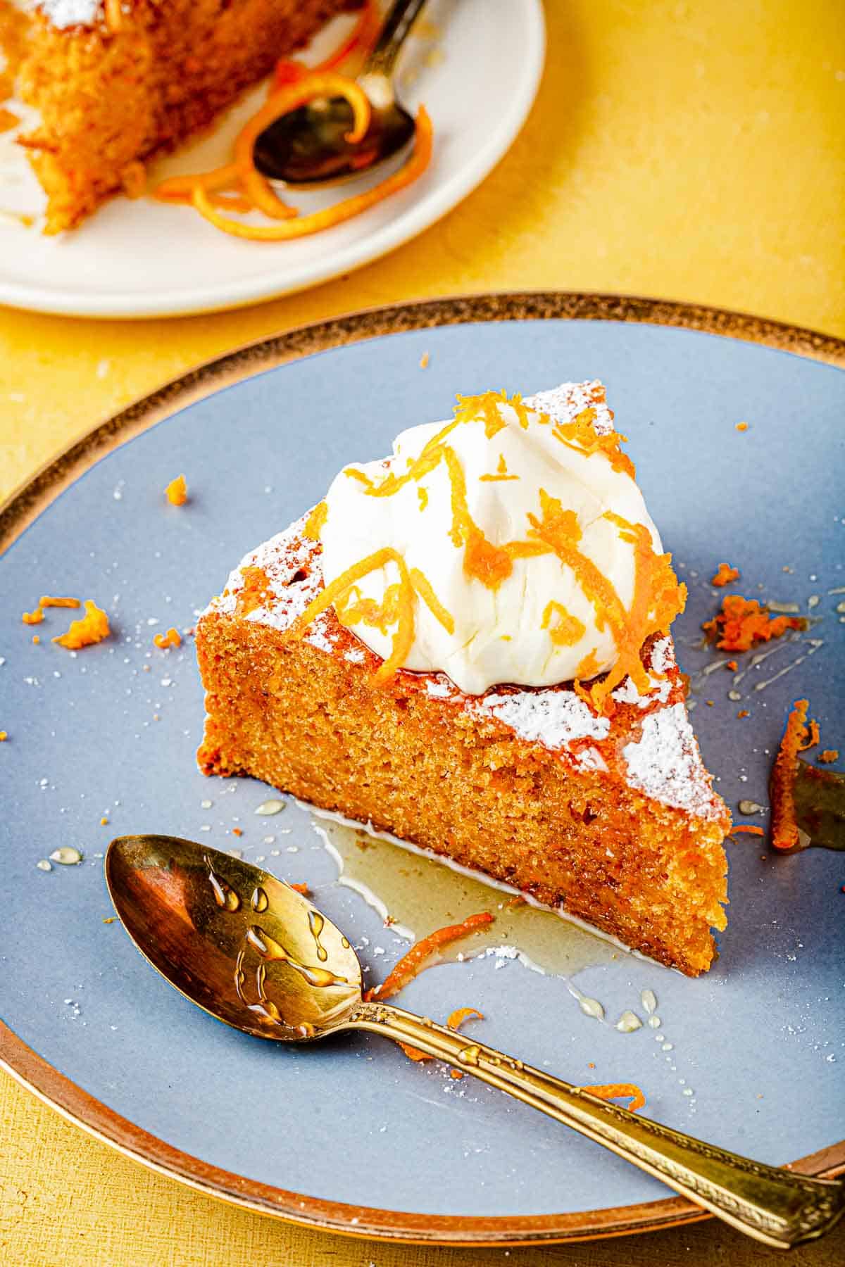 A slice of Torta di Carote (Italian Carrot Cake) topped with a dollop of Greek yogurt, a drizzle of honey and orange zest on a plate with a spoon.