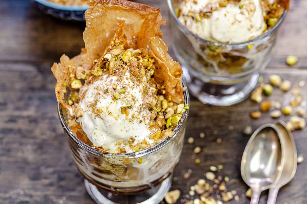close up of a baklava ice cream sundae in a bowl next to another sundae and two spoons..