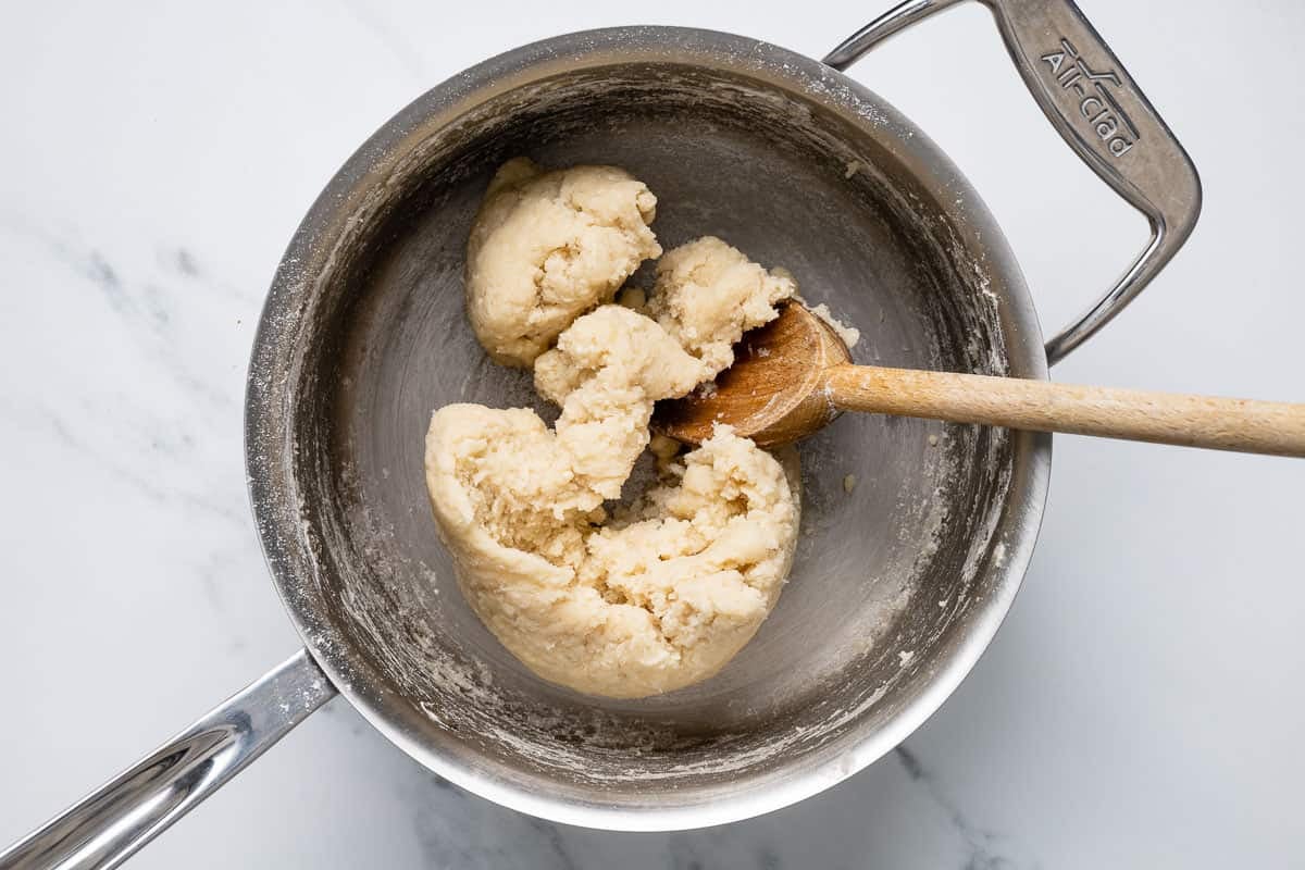 Churro dough being mixed with a wooden spoon in an all-clad pot.