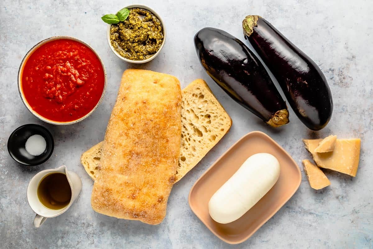ingredients for eggplant parmesan sandwiches including olive oil, ciabatta bread, eggplant, parmesan cheese, mozzarella cheese, and pesto.