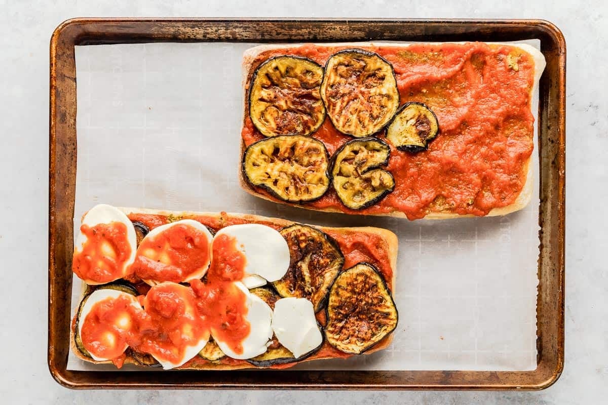 two eggplant parmesan sandwiches being assembled with marinara, eggplant slices and mozzarella on a parchment-line sheet pan.