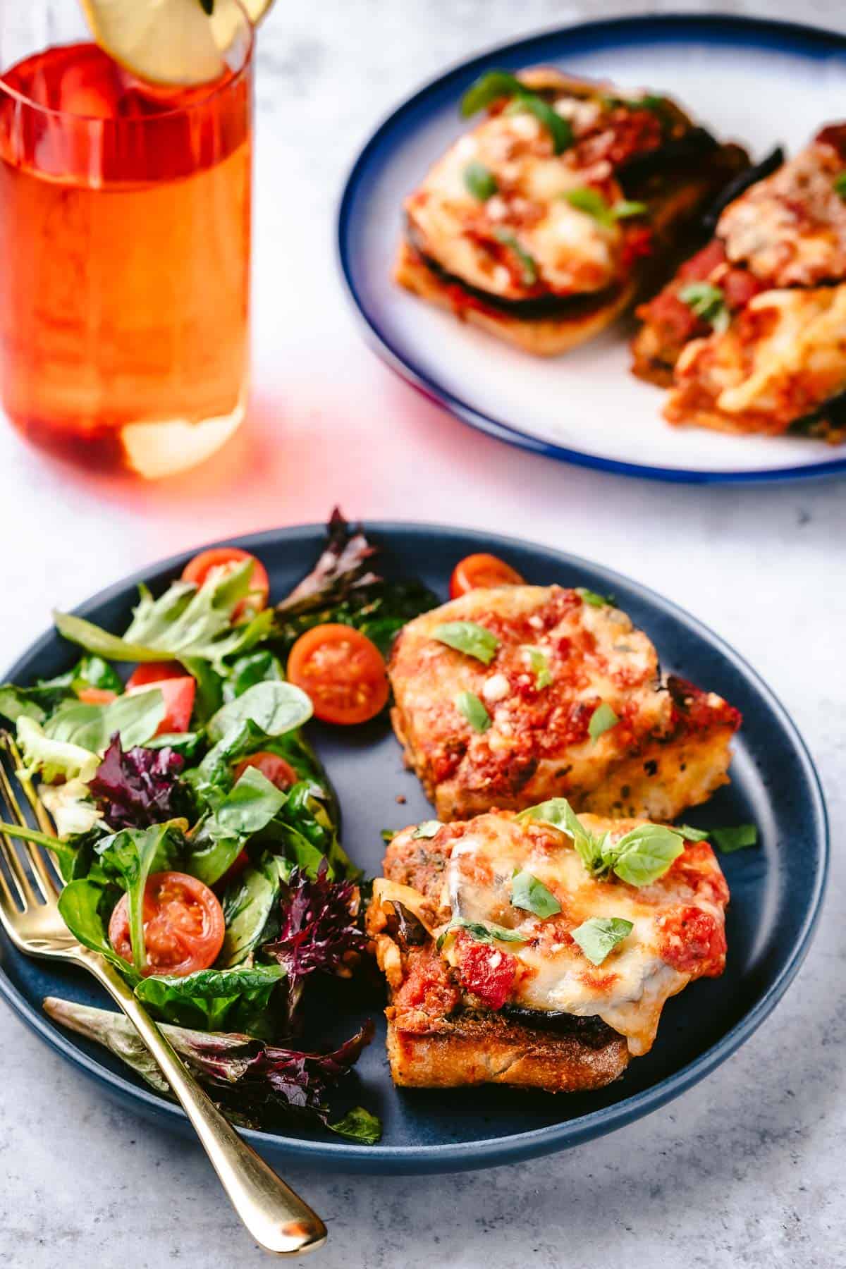 two slices of the eggplant parmesan sandwich on a plate with a salad and a fork next to a glass of tea and another plate with two slices of the eggplant parmesan sandwich.